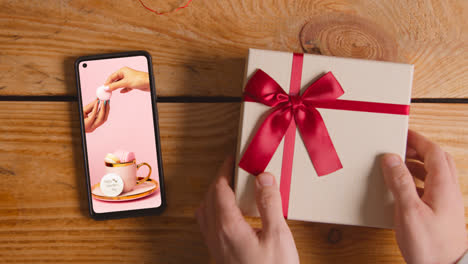 Close-Up-Of-Hand-Picking-Up-Valentine's-Day-Gift-Next-To-Mobile-Phone-With-Valentines-Message-On-Screen